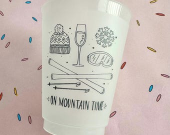 Puede incluir: Vaso de plástico blanco con dibujos lineales negros de equipos de deportes de invierno, incluyendo esquís, gafas y un gorro. El vaso también presenta una copa de champán y un diseño de copo de nieve, con el texto "ON MOUNTAIN TIME".