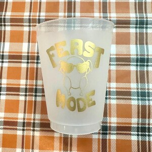May include: A white plastic cup with a gold design that says "FEAST MODE" and a cartoon turkey wearing sunglasses.