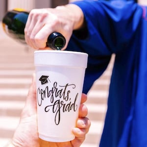 May include: A white foam cup with the text "Congrats Grad" and a graduation cap icon is being filled with champagne.