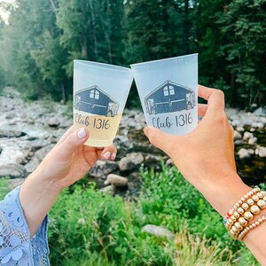 May include: Two clear plastic cups with black text and graphics. The text on the cups reads "Club 1316". The cups are being held by two people.