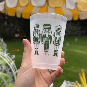 May include: A clear plastic cup featuring three green nutcracker soldier illustrations. The cup is held in front of a yellow and white striped umbrella and a green lawn.