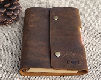 May include: A brown leather-bound journal with a flap closure and a metal snap button. The journal has a decorative mountain and heart design with the text "I ♥ U" embossed on the front cover.