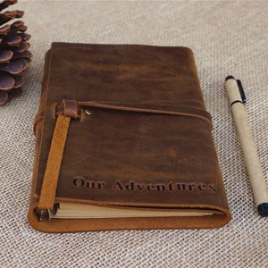 May include: A brown leather journal with a strap closure and the words "Our Adventures" embossed on the front. The journal is lying on a burlap surface with a pen next to it.