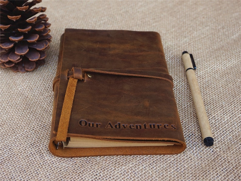 May include: A brown leather journal with a strap closure and the words "Our Adventures" embossed on the front. The journal is lying on a burlap surface with a pen next to it.