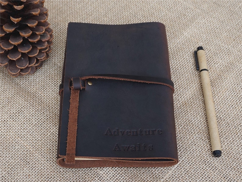 May include: A dark brown leather journal with a strap closure and the words "Adventure Awaits" embossed on the front.