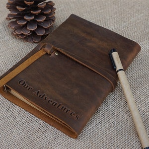 May include: A brown leather journal with a strap closure and the words "Our Adventures" embossed on the front. A brown pen is lying next to the journal.