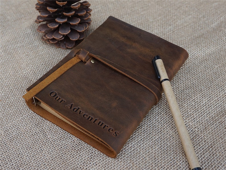 May include: A brown leather journal with a strap closure and the words "Our Adventures" embossed on the front. A brown pen is lying next to the journal.
