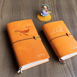 Puede incluir: Dos diarios de cuero naranja con diseños en relieve. Uno presenta una ballena, el otro montañas. Cada diario tiene un cierre elástico negro y un pequeño cierre de lengüeta. Un diario está etiquetado como "Abby". Una pequeña figura de sirena está cerca.