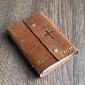May include: A brown leather journal with a cross design and the name "Taylor" embossed on the cover. The journal has a flap closure with two metal snap buttons. The leather has a textured appearance.