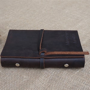 May include: A brown leather journal with a strap closure and the words "Adventure awaits" embossed on the cover. The journal is closed and lying on a hessian surface.