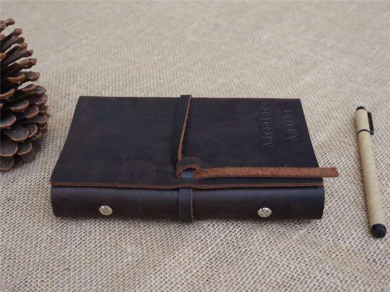 May include: A brown leather journal with a strap closure and the words "Adventure awaits" embossed on the cover. The journal is closed and lying on a hessian surface.