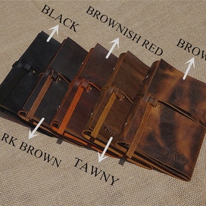 May include: Five leather-bound journals in different shades of brown, black, and reddish brown. The journals have a strap closure and are arranged in a row with the darkest journal on the left and the lightest on the right. The journals are labeled with their colors: Black, Dark Brown, Tawny, Brownish Red, and Brown.