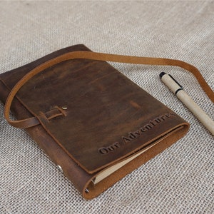 May include: A brown leather journal with a strap closure and the words "Our Adventures" embossed on the cover. A pen is lying next to the journal.