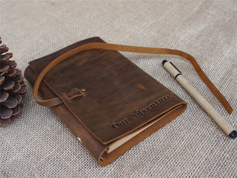 May include: A brown leather journal with a strap closure and the words "Our Adventures" embossed on the cover. A pen is lying next to the journal.