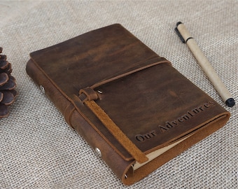 Könnte beinhalten: Ein braunes Lederjournal mit einem Riemenverschluss und den Worten "Our Adventures" geprägt auf dem Cover. Ein brauner Stift liegt neben dem Journal.