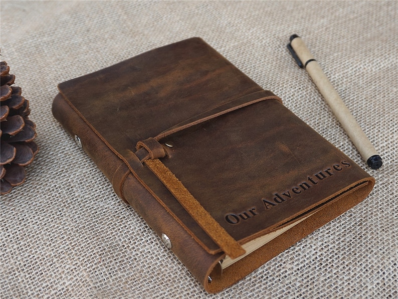 May include: A brown leather journal with a strap closure and the words "Our Adventures" embossed on the cover. A brown pen is lying next to the journal.