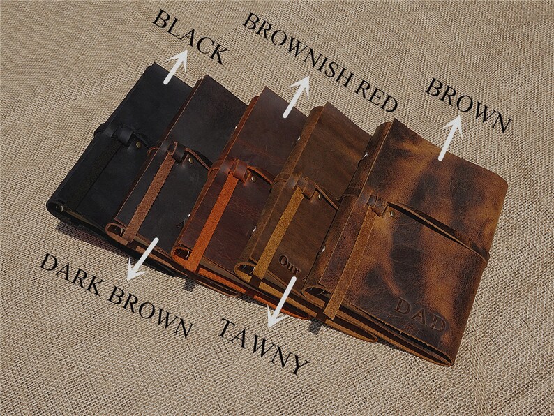 May include: Five leather-bound journals in different shades of brown, black, and reddish brown. The journals have a strap closure and are arranged in a row with the darkest journal on the left and the lightest on the right. The journals are labelled with their colours: Black, Dark Brown, Tawny, Brownish Red, and Brown.