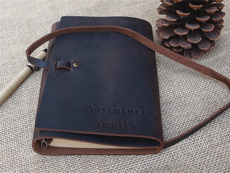 May include: A brown leather journal with a strap closure and the words "Adventure Awaits" embossed on the front.
