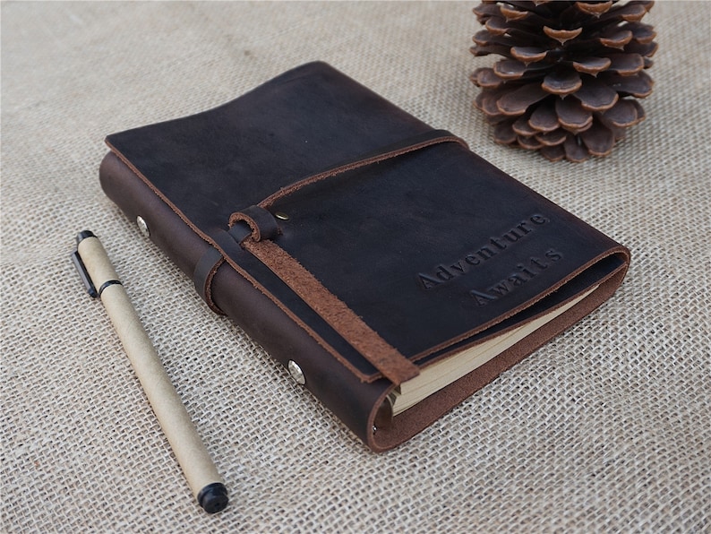 May include: A brown leather journal with a brown leather strap and the words "Adventure Awaits" embossed on the front cover. A brown pen is lying next to the journal.