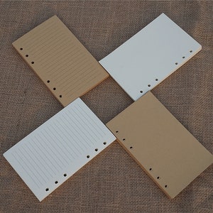 May include: Four sheets of paper with holes punched along the side. Two sheets are brown and two sheets are white. The sheets are lined and are lying on a brown hessian surface.