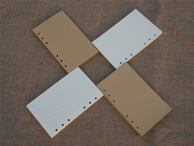 May include: Four sheets of paper with holes punched along the side. Two sheets are brown and two sheets are white. The sheets are lined and are lying on a brown hessian surface.
