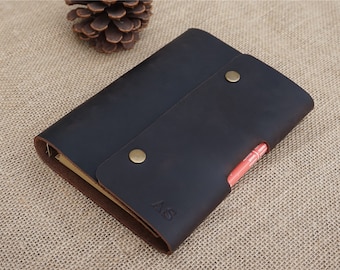 May include: A black leather notebook with a flap closure and a pen holder. The notebook has two brass snaps and the initials "AS" are embossed on the front.