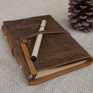 May include: A brown leather journal with a pen tucked inside. The journal has a strap closure and the words "Our Adventures" are embossed on the front cover.