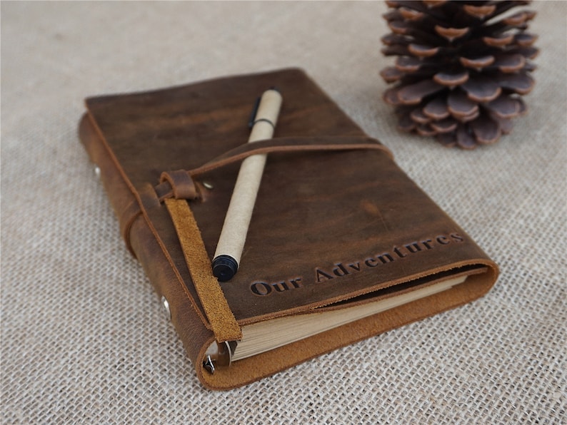 May include: A brown leather journal with a pen tucked inside. The journal has a strap closure and the words "Our Adventures" are embossed on the front cover.