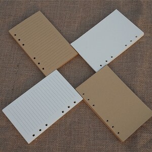 May include: Four sheets of paper with holes punched along the side. Two sheets are brown and two sheets are white. The sheets are lined and are lying on a brown burlap surface.