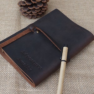 May include: A brown leather journal with a brown leather strap and the words "Adventure Awaits" embossed on the front. A brown pen is lying on top of the journal.