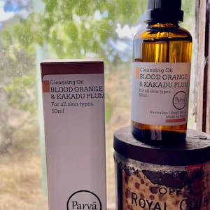 May include: Two bottles of cleansing oil labeled "Blood Orange & Kakadu Plum" by Parva. The bottles are 50 ml and are made in Australia. The bottles are sitting on a wooden surface next to a vintage tin with the text "Cope's Royal Club" on it.