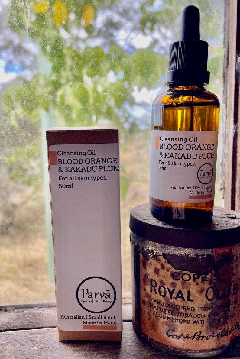 May include: Two bottles of cleansing oil labeled "Blood Orange & Kakadu Plum" by Parva. The bottles are 50 ml and are made in Australia. The bottles are sitting on a wooden surface next to a vintage tin with the text "Cope's Royal Club" on it.