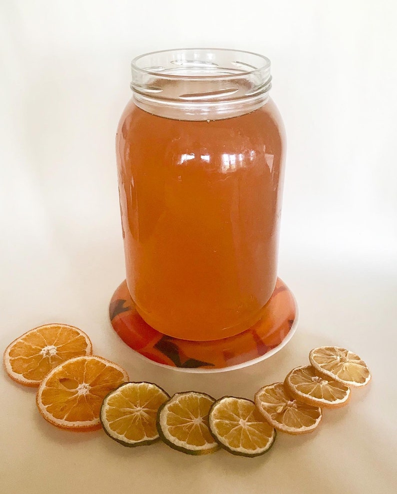 May include: A glass jar filled with a light orange liquid, sitting on a round orange and white coaster. Dried orange and lime slices are arranged in a semi-circle in front of the jar.