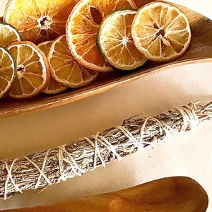 May include: A wooden bowl filled with dried orange and lime slices. A bundle of sage is tied with twine and a wooden spoon is on a white surface.