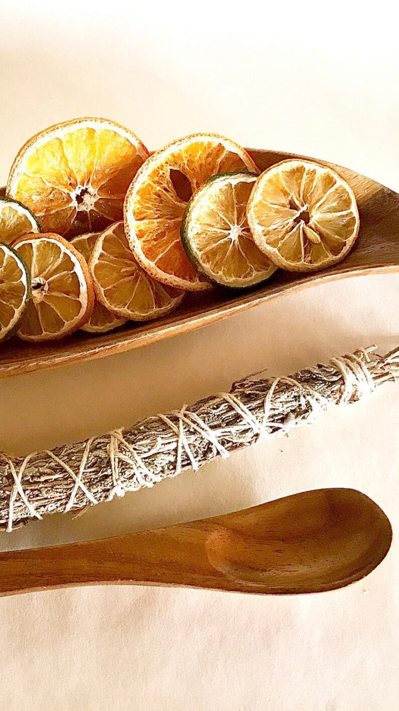 May include: A wooden bowl filled with dried orange and lime slices. A bundle of sage is tied with twine and a wooden spoon is on a white surface.