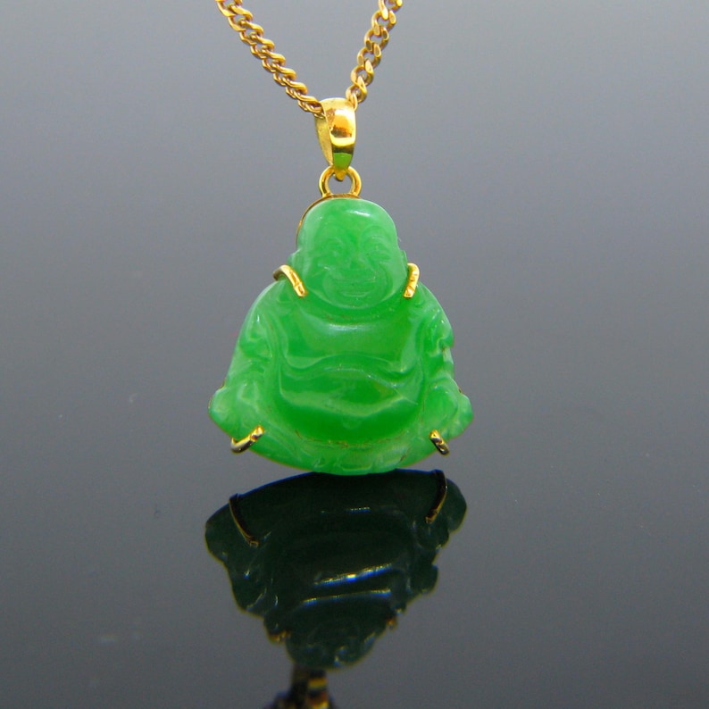 May include: A green jade pendant of a smiling Buddha figure, carved in a sitting pose, with gold accents around the edges. The pendant is attached to a gold chain.