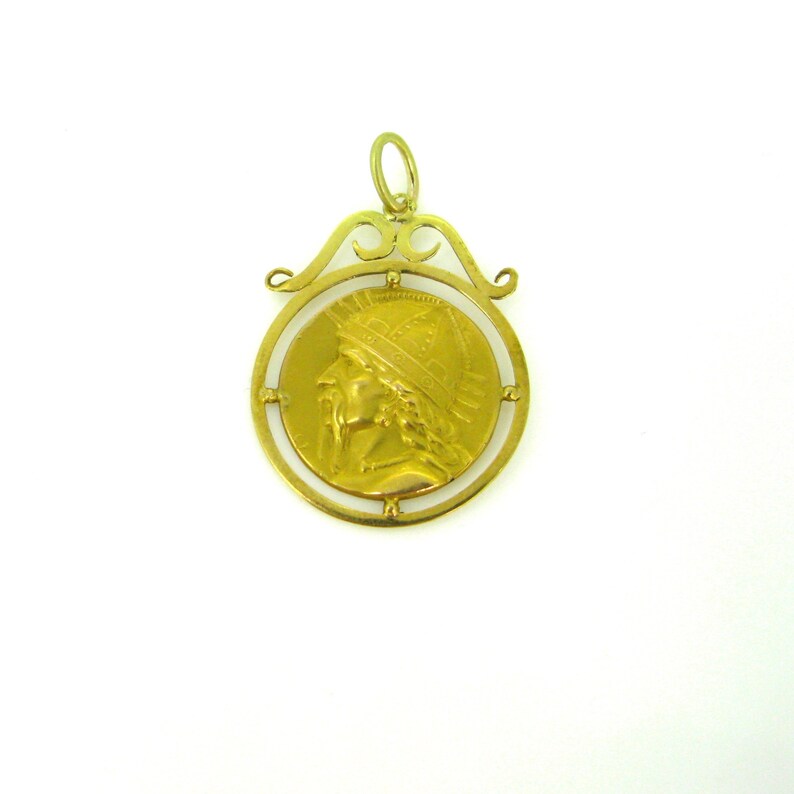 May include: A gold-colored pendant featuring a circular coin design with a profile of a person wearing a helmet. The coin is set within a decorative frame with an ornate bail for hanging. The pendant is on a white background.