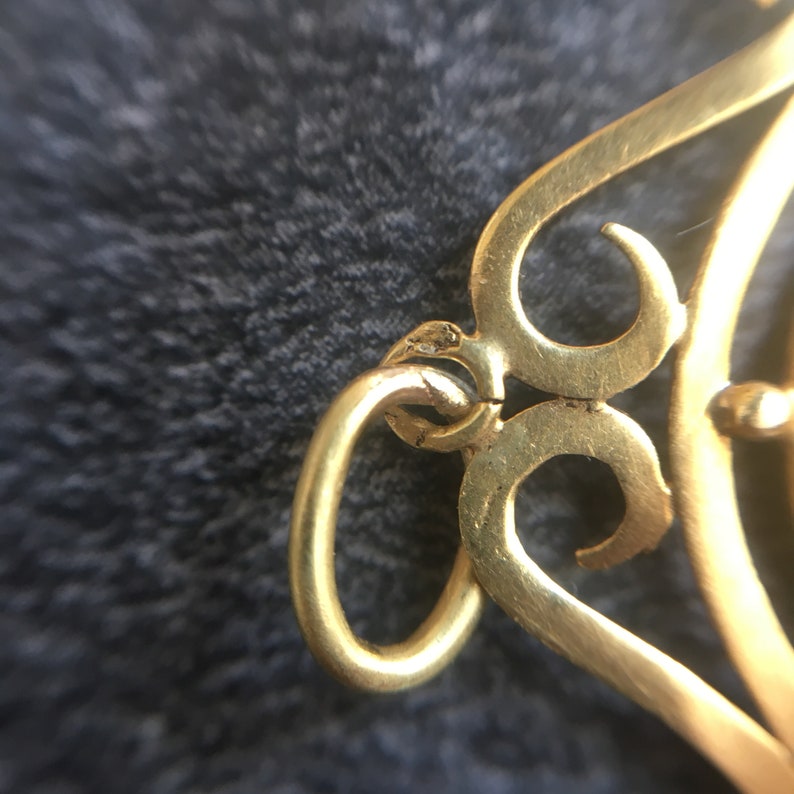 May include: Close-up of a gold-colored jewelry piece with a decorative, openwork design. The piece features a circular loop and intricate, swirling patterns. The metal has a slightly aged appearance.