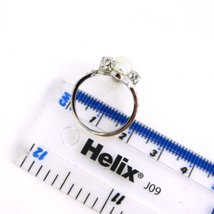 May include: Silver ring with two white pearls and clear rhinestones. The ring is measured on a ruler with the Helix brand name and J09 printed on it.