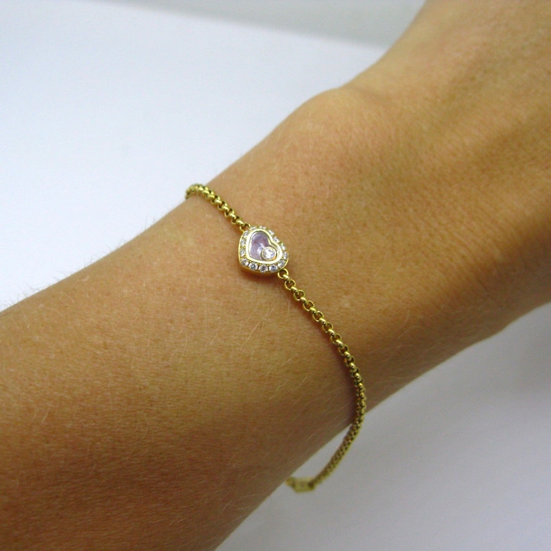 May include: A gold chain bracelet with a heart-shaped pendant featuring a small pink stone surrounded by clear stones.