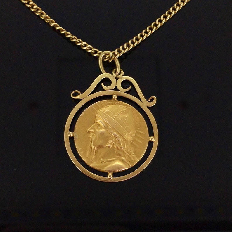 May include: A gold-colored pendant necklace featuring a circular coin design with a profile of a person wearing a crown and a long beard. The pendant is suspended from a delicate gold chain. The necklace is displayed against a dark background.
