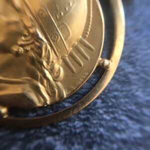 May include: Close-up of a gold-colored pendant with a detailed relief design. The pendant features a classical figure and intricate details, set within a circular frame. The surface has a polished finish, reflecting light.