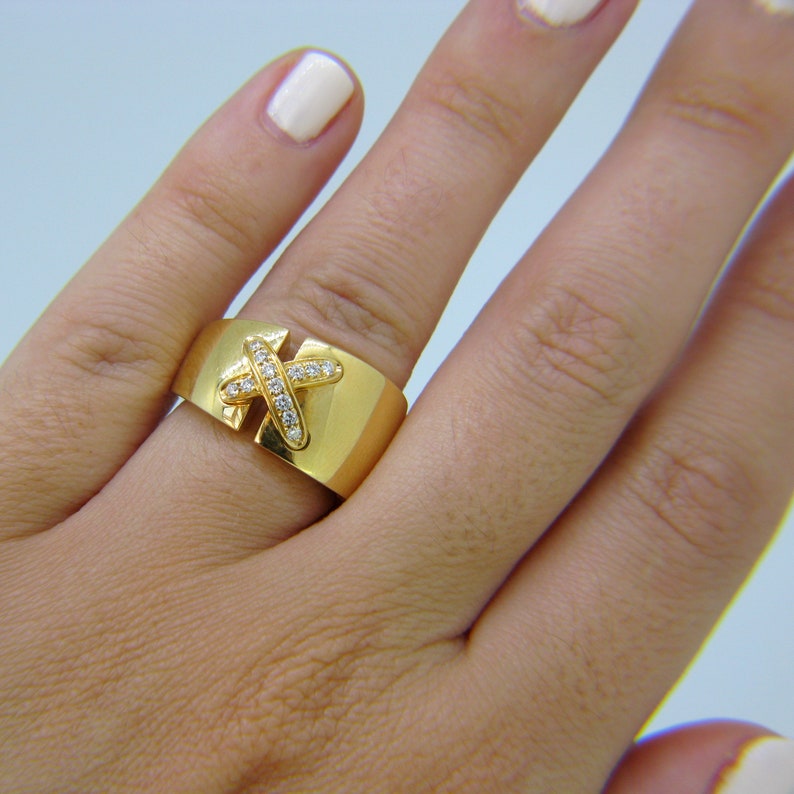 May include: A gold ring with a diamond-encrusted X design. The ring is wide and has a smooth, polished finish.