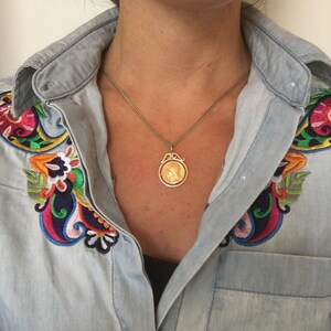 May include: A gold-toned pendant necklace with a detailed circular design. The pendant hangs from a delicate chain. The model is wearing a light blue denim shirt with colorful embroidered floral designs on the shoulders.