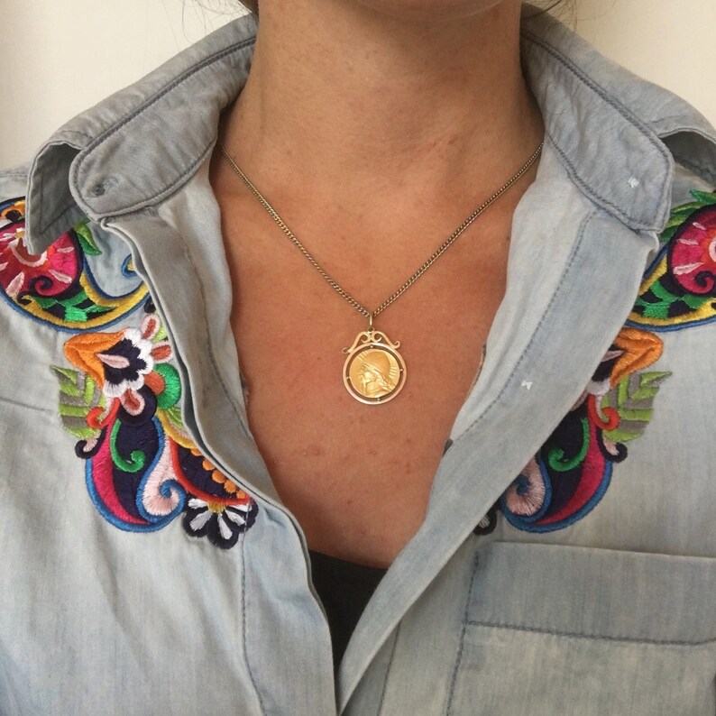 May include: A gold-toned pendant necklace with a detailed circular design. The pendant hangs from a delicate chain. The model is wearing a light blue denim shirt with colorful embroidered floral designs on the shoulders.