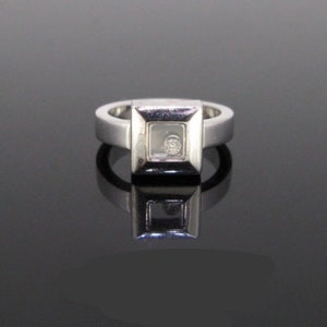 May include: A silver ring with a square-shaped setting that contains a floating diamond. The diamond is visible through a clear glass window.