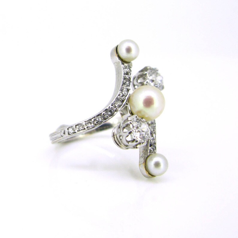 May include: A silver ring with a curved band set with small diamonds and three pearls. The ring has a unique design with the pearls set in a cluster.