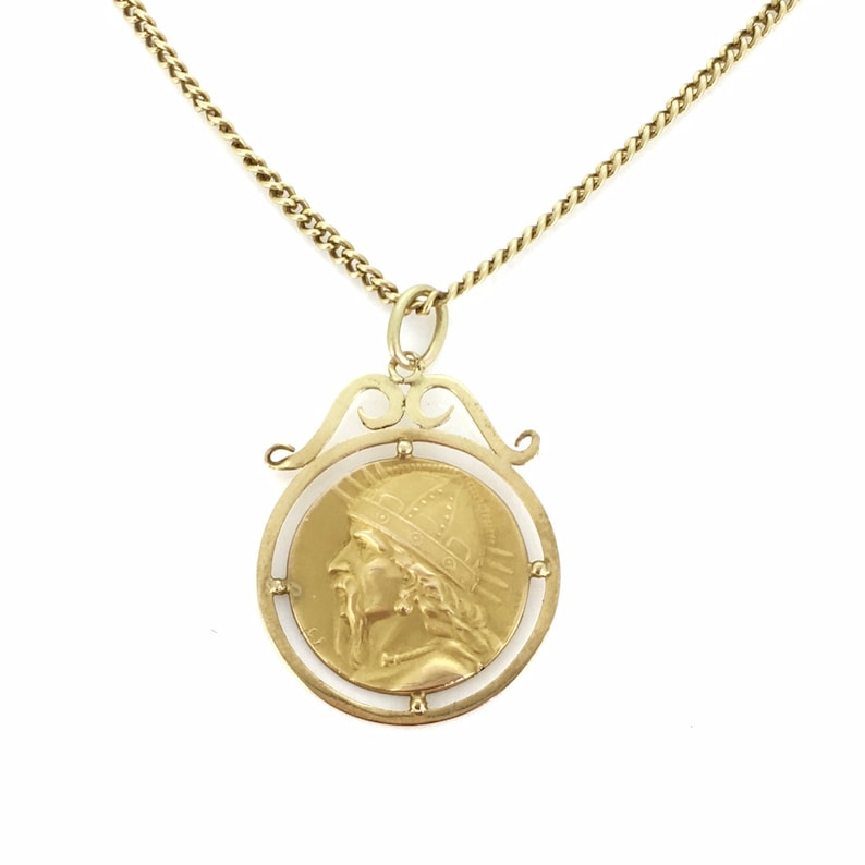 May include: A gold-toned necklace featuring a circular pendant with a profile of a man wearing a helmet. The pendant is suspended from a delicate chain. The pendant has an ornate frame and is likely a vintage or antique piece.