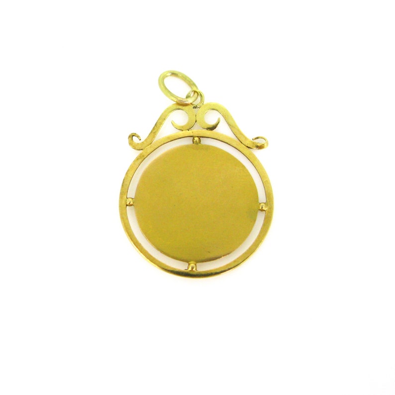 May include: A gold-colored pendant with a circular design. The pendant has a decorative top with a loop for a chain. The center of the pendant is a solid gold color, and the outer frame is a thin gold outline.