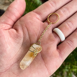 Op de afbeelding: Een sleutelhanger met een gouden ketting en een helder kristal dat is gewikkeld in gouden draad. Het kristal is lichtgeel.